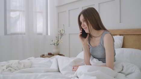 Young-caucasian-woman-sitting-on-the-bed-in-the-morning-feels-bad.-A-young-woman-is-making-a-smartphone-call-to-her-doctor.